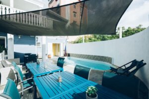 Terrasse extérieure avec des tables bleues en bois située au bord d'une piscine sous une voile d'ombrage.
