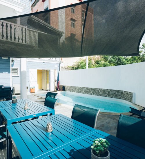 Terrasse extérieure avec des tables bleues en bois située au bord d'une piscine sous une voile d'ombrage.