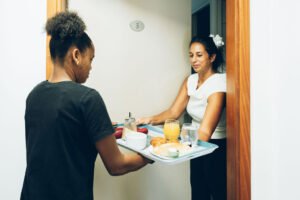 Une employée tend un plateau repas garni à une cliente à l'entrée d'une chambre d'hôte