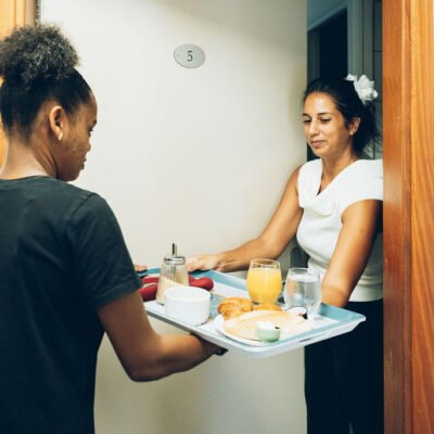 Une employée tend un plateau repas garni à une cliente à l'entrée d'une chambre d'hôte