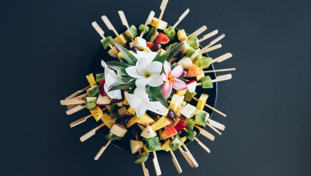 Vue de dessus d'un plat de brochettes de fruits frais disposées en cercle et décorées de fleurs de frangipanier.