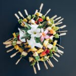 Vue de dessus d'un plat de brochettes de fruits frais disposées en cercle et décorées de fleurs de frangipanier.