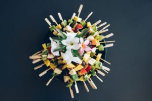 Vue de dessus d'un plat de brochettes de fruits frais disposées en cercle et décorées de fleurs de frangipanier.